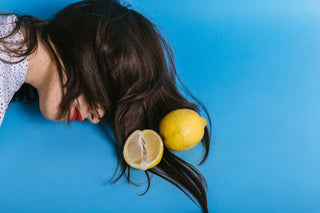 On a bright blue background a woman lies with her brown hair covering her eyes. She has two lemons in her hair forming a bright colour contrast with the background.