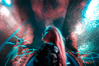 A woman with long pink air stands in an aquarium. the lighting around her is pink and light blue and makes for a sci-fi feel.