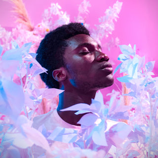A dark skinned young man has his eyes closed in a sea of neon lilac flowers in front of a pink background.