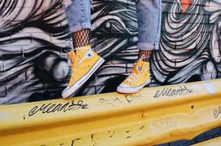in front of a colourful mural, a set of legs wearing jeans with visible fishnets and bight yellow converse high-tops walks across a yellow bollard. 