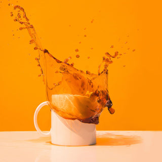In front of an orange background, a white mug sits on a white bench. The moment captured shows black coffee exploding from the mug, as if someone has just dropped something in the cup.