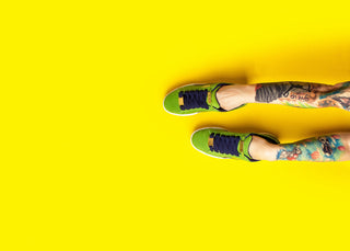 On a bright yellow background sits a pair of colourfully tattoo-ed legs wearing bright green sneakers.