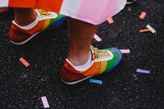 A pair of rainbow-sneakered feed stands on pavement with colourful confetti around the. The tip of a flag can be seen at the edge of the image.