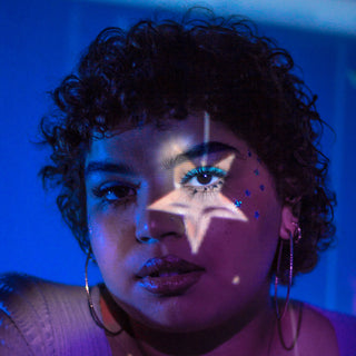 A woman with short curly hair sits in front of a dark blue background. A light colour projection of a star appears over one of her eyes.