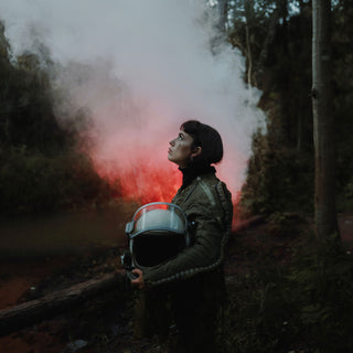 A woman holds a helmet and looks upwards in a forest, red coloured smoke rising up behind her.