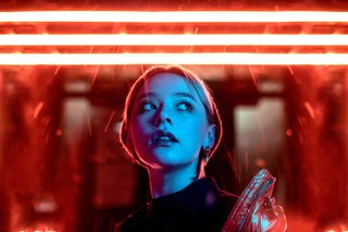 A woman looks forward, bright blue light illuminating her face. Above her, three bands of bright neon red light cast the background in a red vibe.