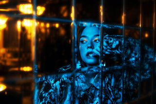 Standing behind a wire fence is a young woman with shiny eye-shadow. While the background is warm yellow lighting, she has a blue glow in front of her.