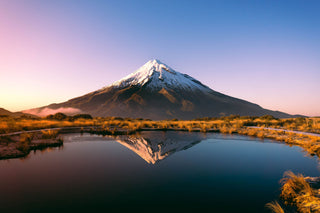 A mountain in Auckland New Zealand sits in front of a soft setting sky. The mountain is reflected in a pool of still water at the base of the image.