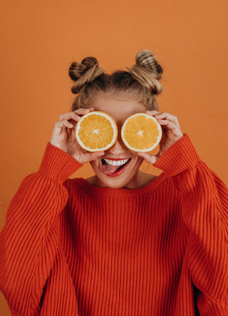 A woman with hair in space buns stands in front of an orange background and wears a bright orange jumper. She holds orange halves in front of her eyes and has her tongue sticking out.